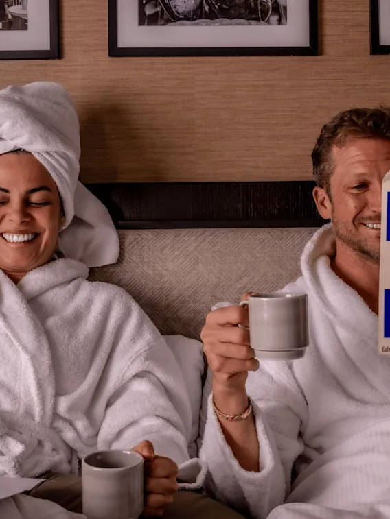 A man and a woman wearing white bathrobes relaxing in a hotel bed. The woman has a towel wrapped around her head and is laughing, while the man holds a mug and reads a book titled "Intermezzo" by Sally Rooney.