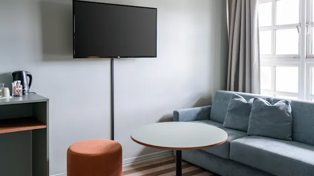 En veggmontert TV er av i et minimalistisk rom med en grønn sofa, rundt bord og oransje krakk. Mykt naturlig lys kommer inn gjennom et gardinbekledd vindu.