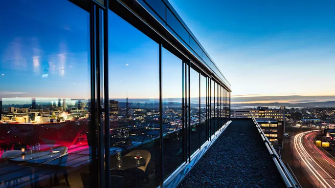 Glass-walled rooftop restaurant reflects the night city skyline, with interior tables dimly lit. Adjacent road below shows light trails from moving vehicles.