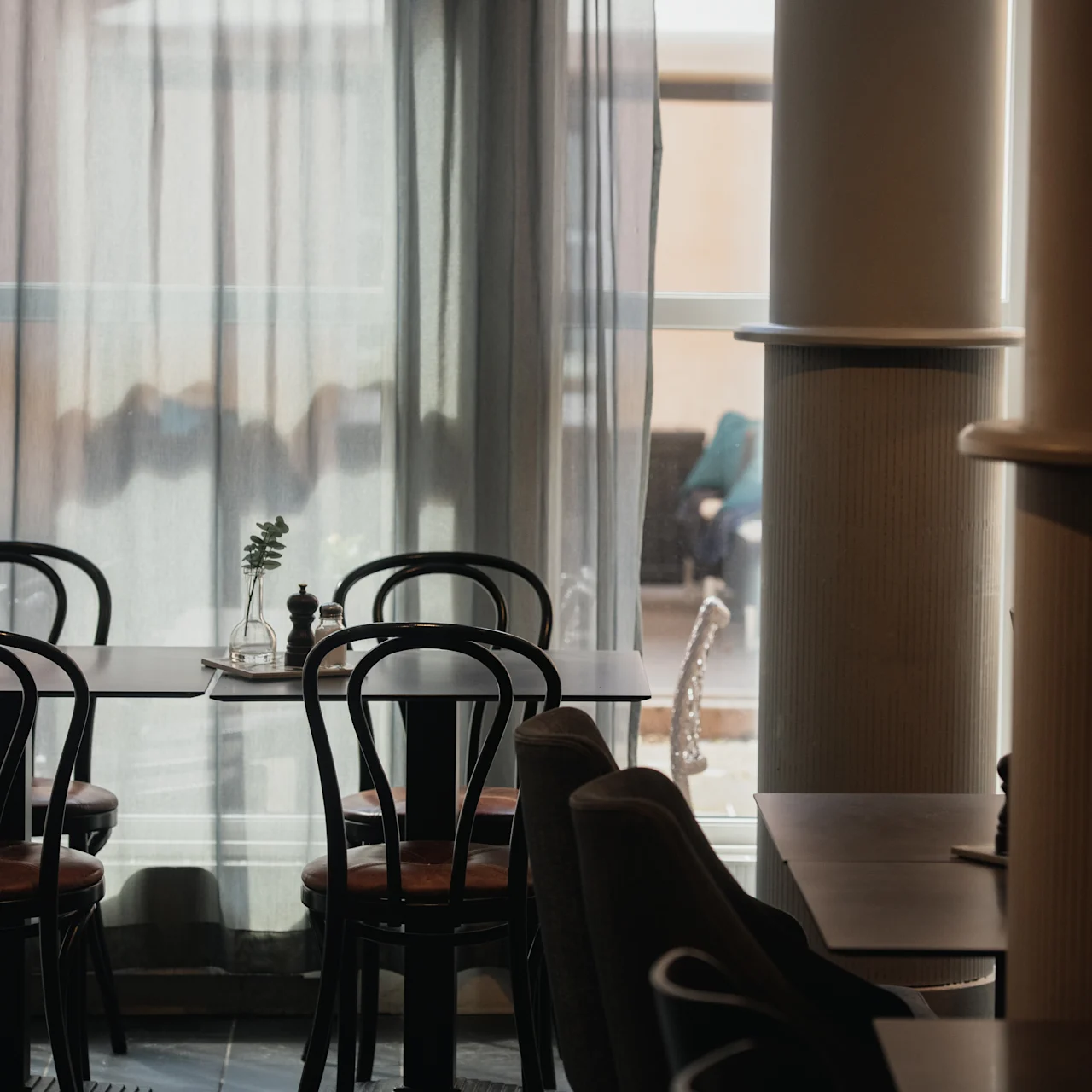 Four black chairs are placed around a wooden table with a small vase, in a softly lit dining area near large windows with sheer curtains.