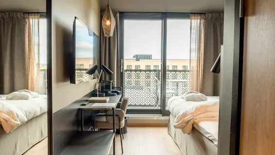 Ett modernt hotellrum med säng, skrivbord, stol och platt-TV som leder till en balkong med utsikt över staden.