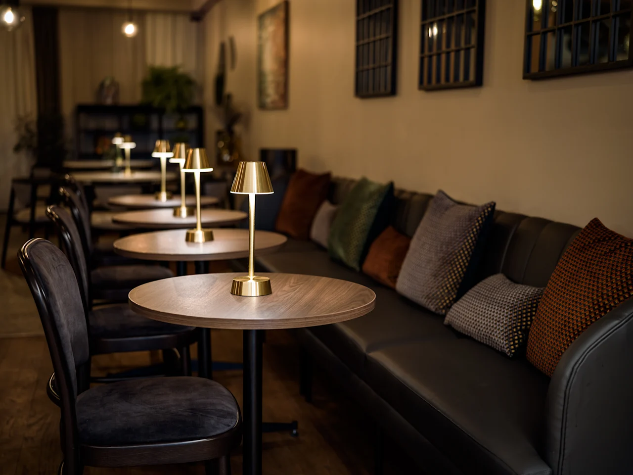 Gyllene bordslampor lyser upp runda trämöbler i ett mysigt, svagt belyst café med vadderade sittplatser. Mjuka kuddar pryder bänkar mot de varma väggarna, vilket skapar en inbjudande atmosfär.
