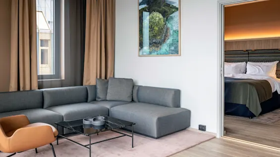 En grå hjørnesofa står på et lyserødt tæppe ved siden af en læderstol og sort sofabord; gardiner dækker et vindue. Et soveværelse med en redt seng ses gennem en åben dør.