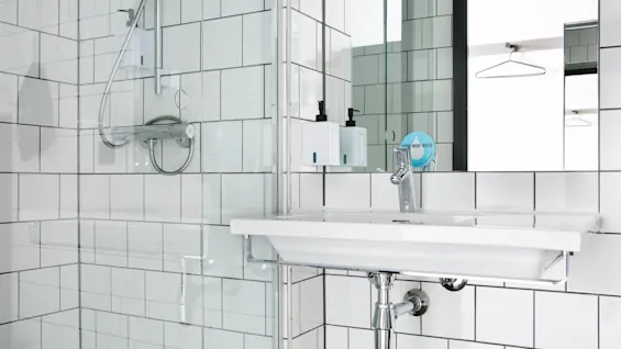 Handfat monterat i ett kaklat badrum med kromkran och tvåldispensrar. Duscharmatur till sidan. Speglar reflekterar en hängande galge. Text: "DRINK MORE WATER".