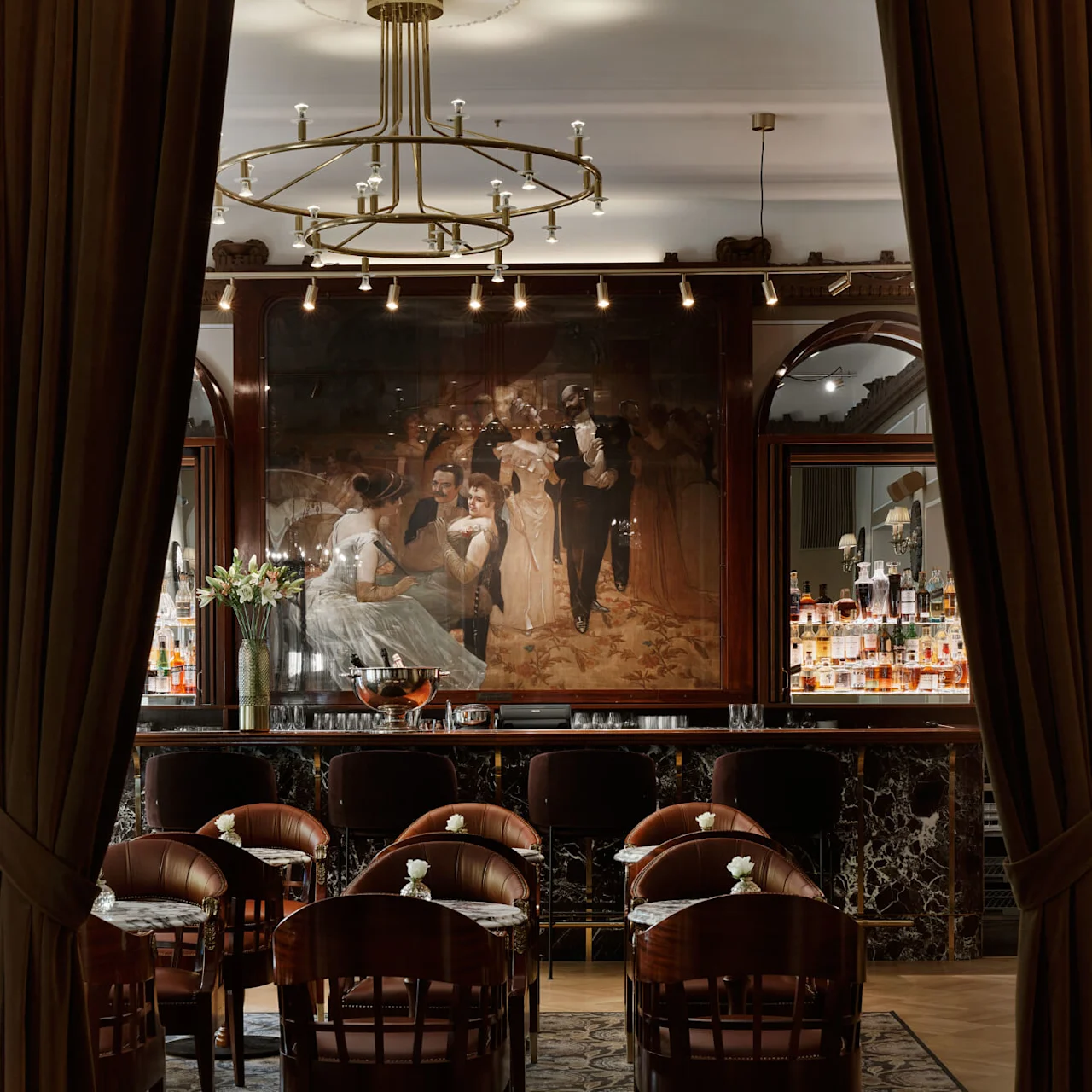 Elegant bar area with high-backed chairs, a chandelier overhead, and a large painting reflecting a social scene; plush, classic ambiance at Hotel Kämp in Helsinki.