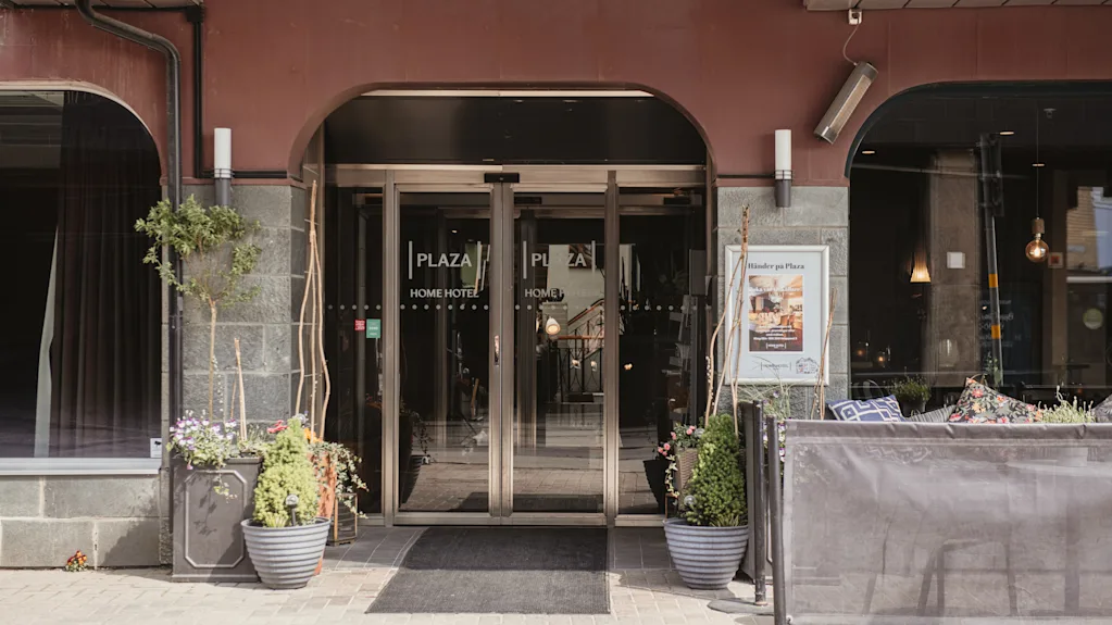Ingången av ett hotell med glasdörrar och krukväxter, omgiven av en röd och grå fasad. Text på dörrarna: "PLAZA HOME HOTEL".