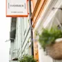 Skilt med teksten "HAMMER HOME HOTEL" henger på en fargerik bygningsfasade. To hengende kurver med grønt pryder forgrunnen. Gaten strekker seg inn i avstanden under en overskyet himmel.