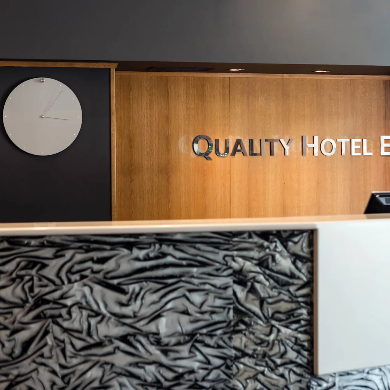 A hotel reception desk with a modern design stands under a wooden wall displaying the text “QUALITY HOTEL ENTRY.” A white clock hangs on a dark backdrop.
