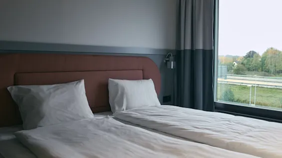 Ett mysigt hotellrum på Stopover Hotel Falkenberg med två sängar och ett stort fönster med utsikt över en väg och grönt landskap.