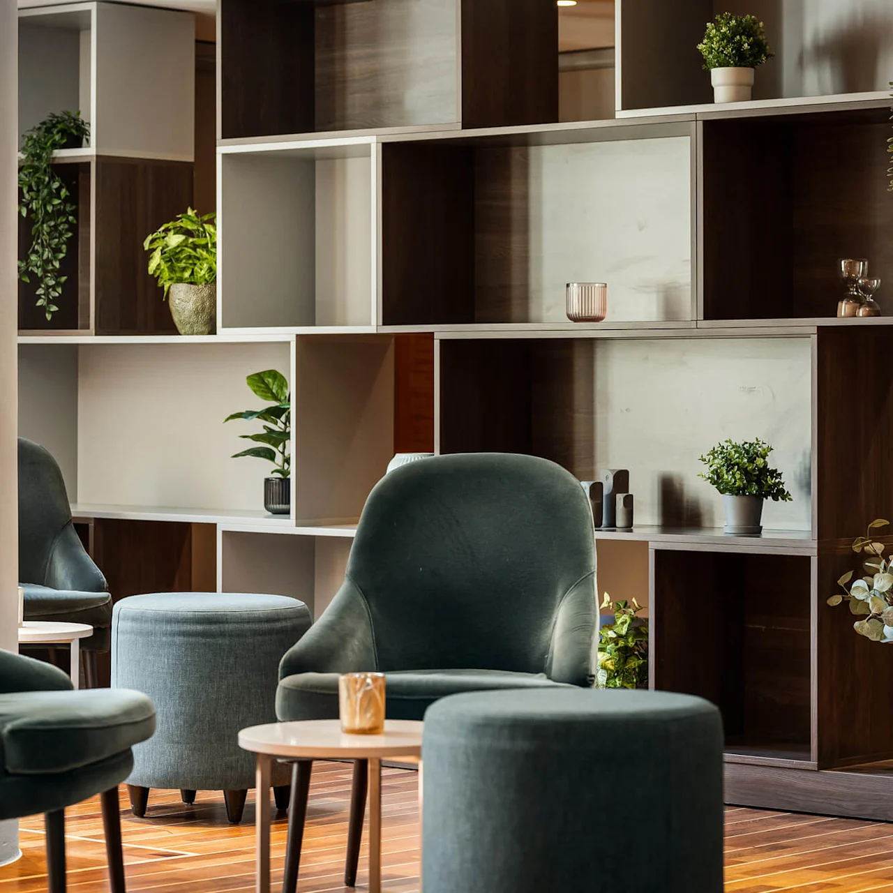 Komfortable stole omgiver små borde i en hyggelig lounge, med planter og dekorative genstande på moderne træhylder mod en lys væg.