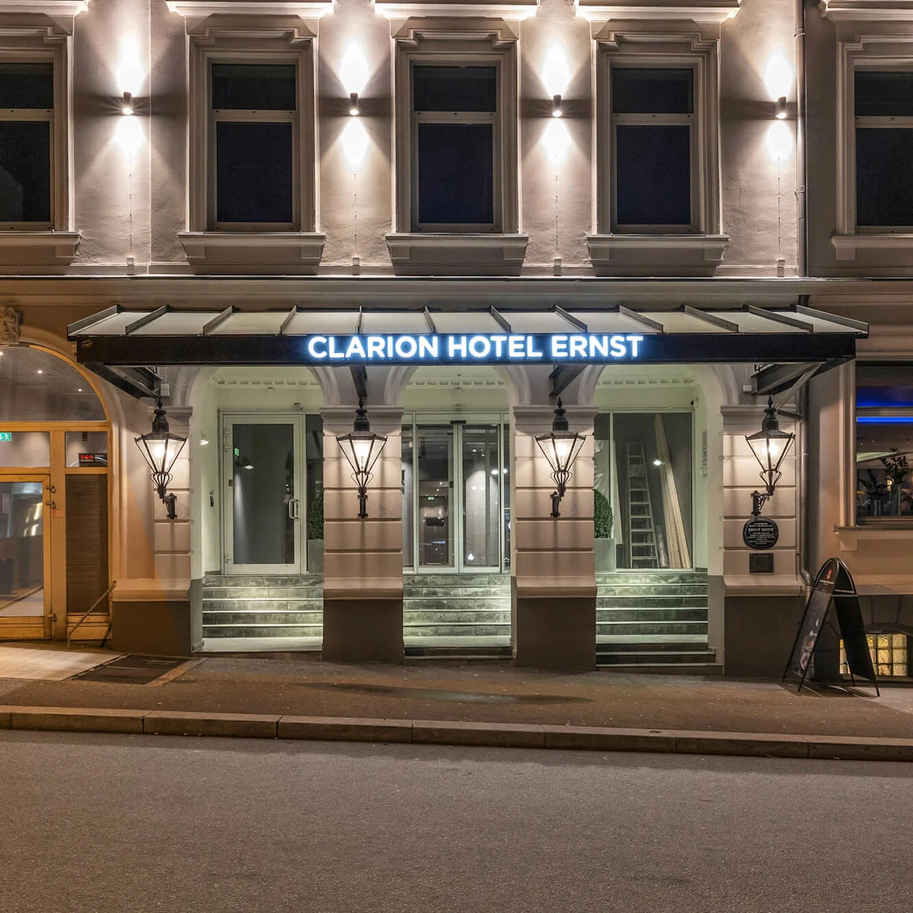 Indgangen til Clarion Hotel Ernst er oplyst om natten, med et skilt og traditionelle lamper på en historisk bygningsfacade. Skiltet lyder "Clarion Hotel Ernst".