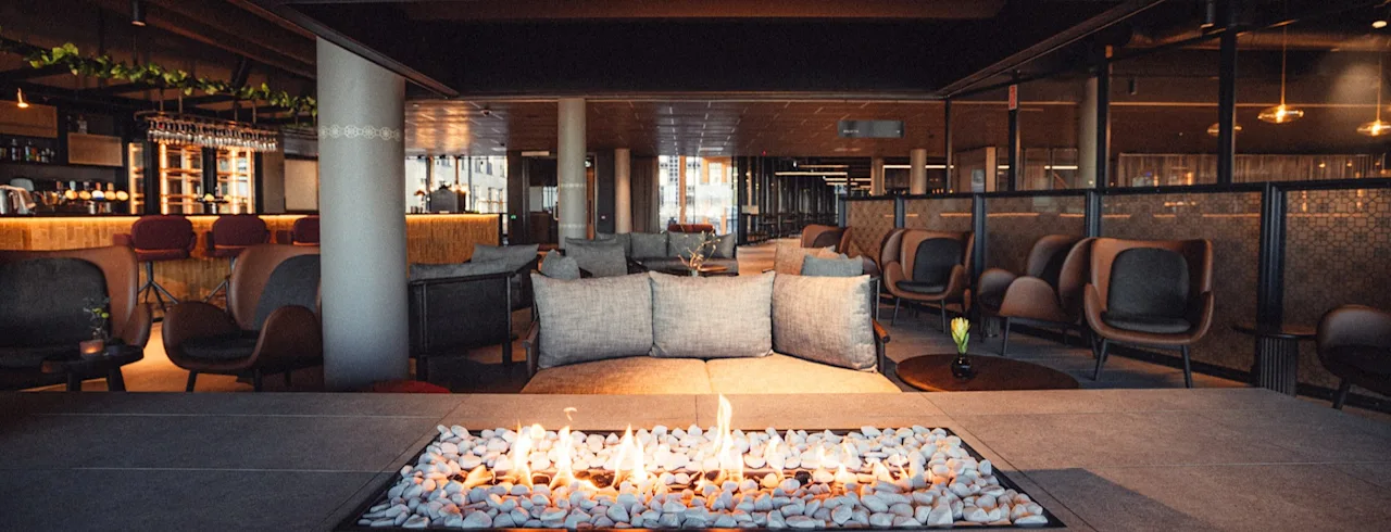 En moderne lounge har en rektangulær ildsted i midten med flammer over sten, omgitt av stoler og sofaer. Miljøet er en stilren innendørs arena med varm belysning.