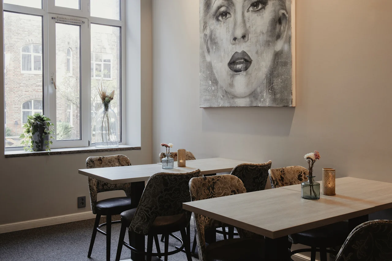 An inviting dining space featuring light wooden tables, patterned chairs, and a large window. A striking black and white portrait adorns the wall.