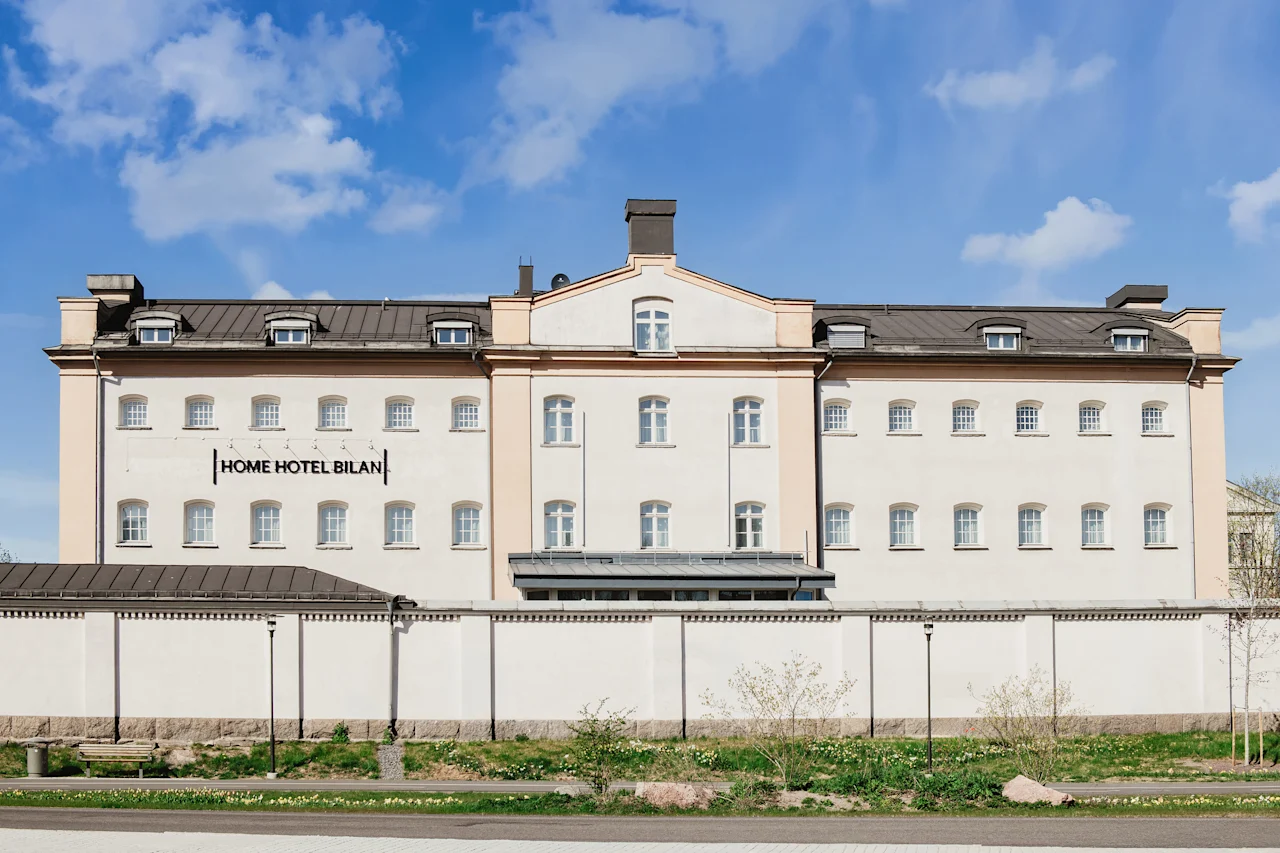 A large, light-colored building with many windows and a dark roof under a blue sky. A white wall stands in front of it. Text on building: HOME HOTEL BILAN.