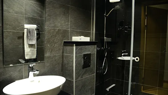 A modern bathroom with dark tiled walls, a white sink, and a glass shower enclosure, suggesting a clean and functional space.