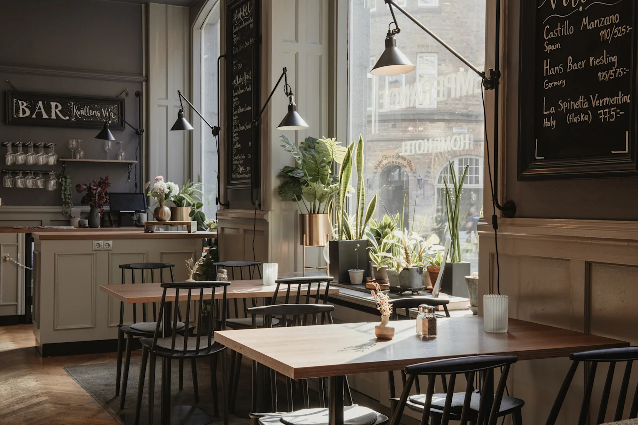 Et koselig bar- eller restaurantinteriør med trebord, stoler og mange planter ved et stort vindu. Et skilt leser BAR Kallens Vi.