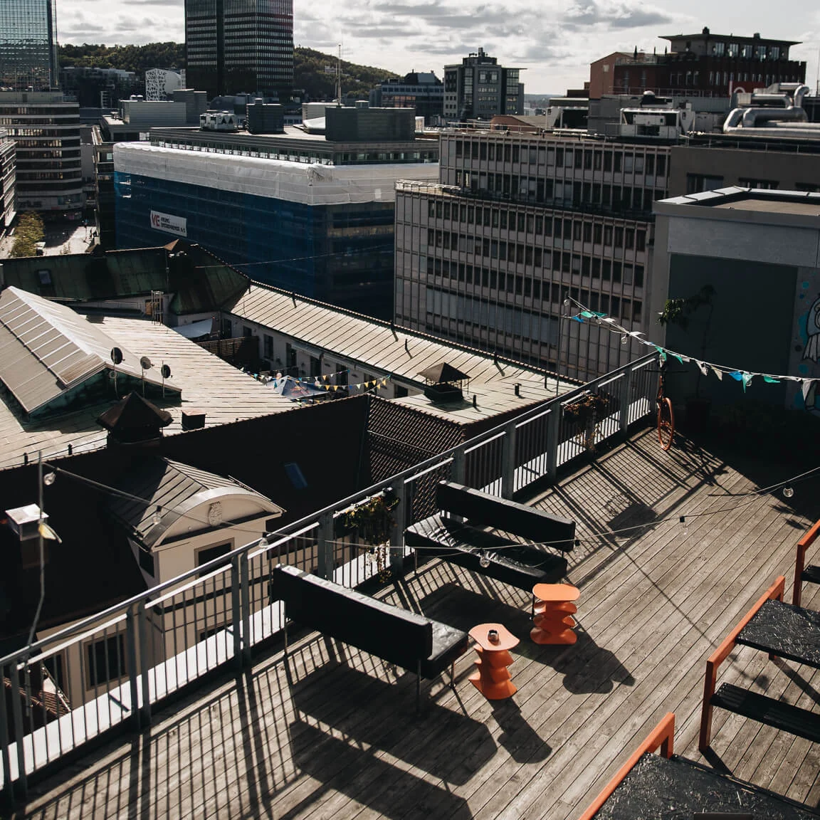 Rooftop terrace with benches and orange tables; a person on a laptop sits at a table. City buildings, hills, and a partly cloudy sky in the background.
