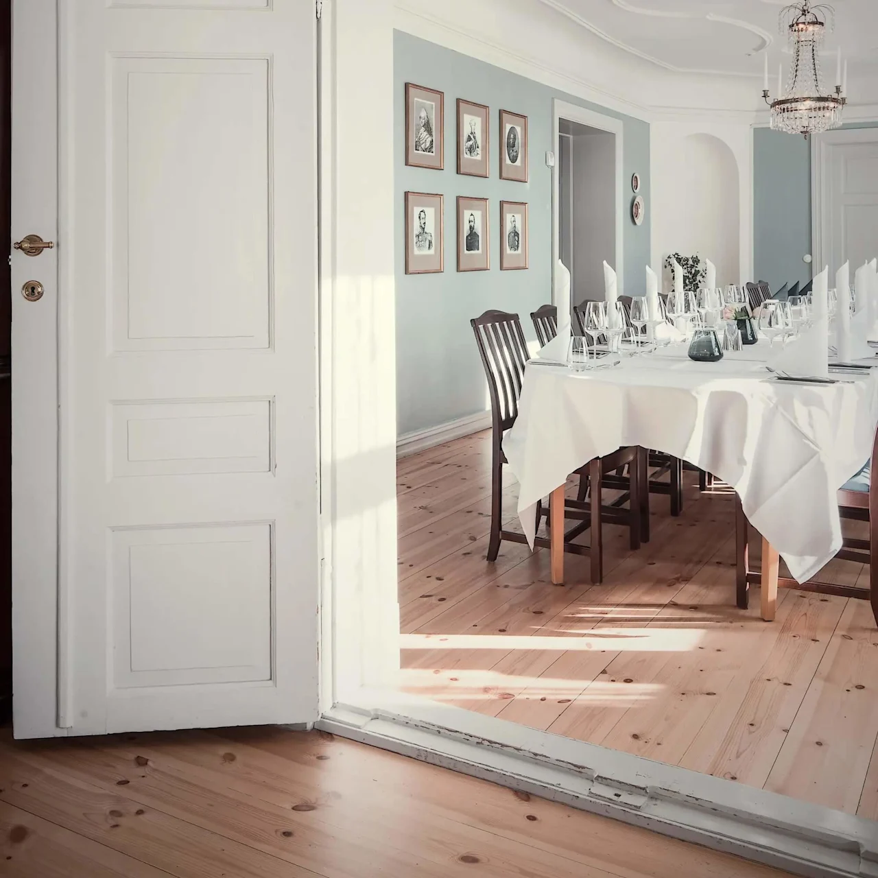 Et elegant dækket bord med hvid dug og servietter, omgivet af træstole, står i et klassisk rum med en lysekrone og indrammede portrætter på væggen.