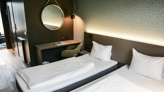Two neatly made single beds stand side by side in a modern hotel room, featuring a decorative wall, a round mirror above a desk, and a soft chair.