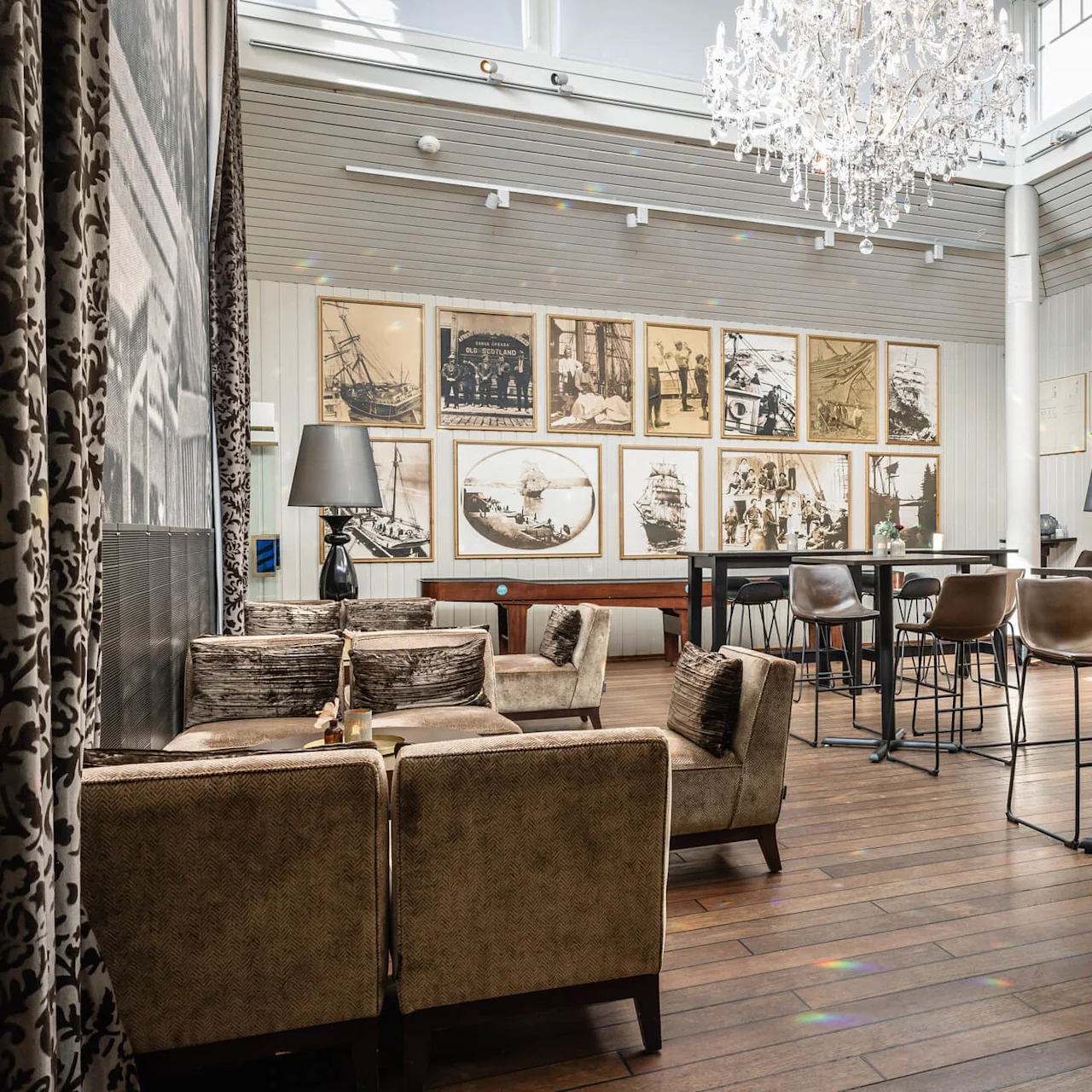 En luksuriøs lysekrone henger over en elegant lounge med vintagefotografier på veggene, brune polstrede stoler rundt bordene. Lys naturlig lys filtreres gjennom store vinduer og skaper en sofistikert, luftig atmosfære.