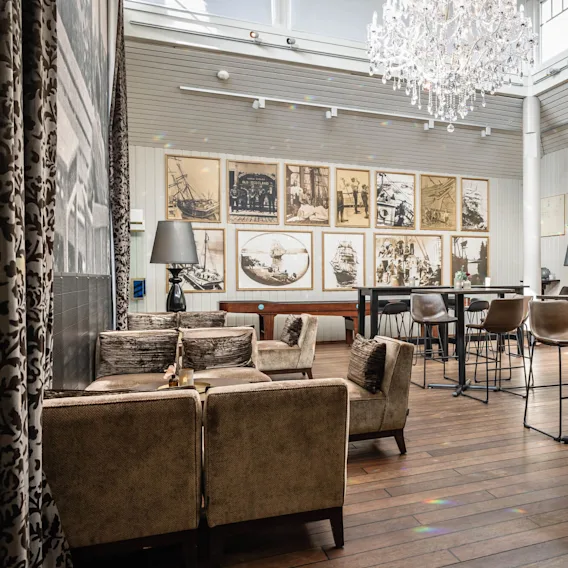 En luksuriøs lysekrone henger over en elegant lounge med vintagefotografier på veggene, brune polstrede stoler rundt bordene. Lys naturlig lys filtreres gjennom store vinduer og skaper en sofistikert, luftig atmosfære.