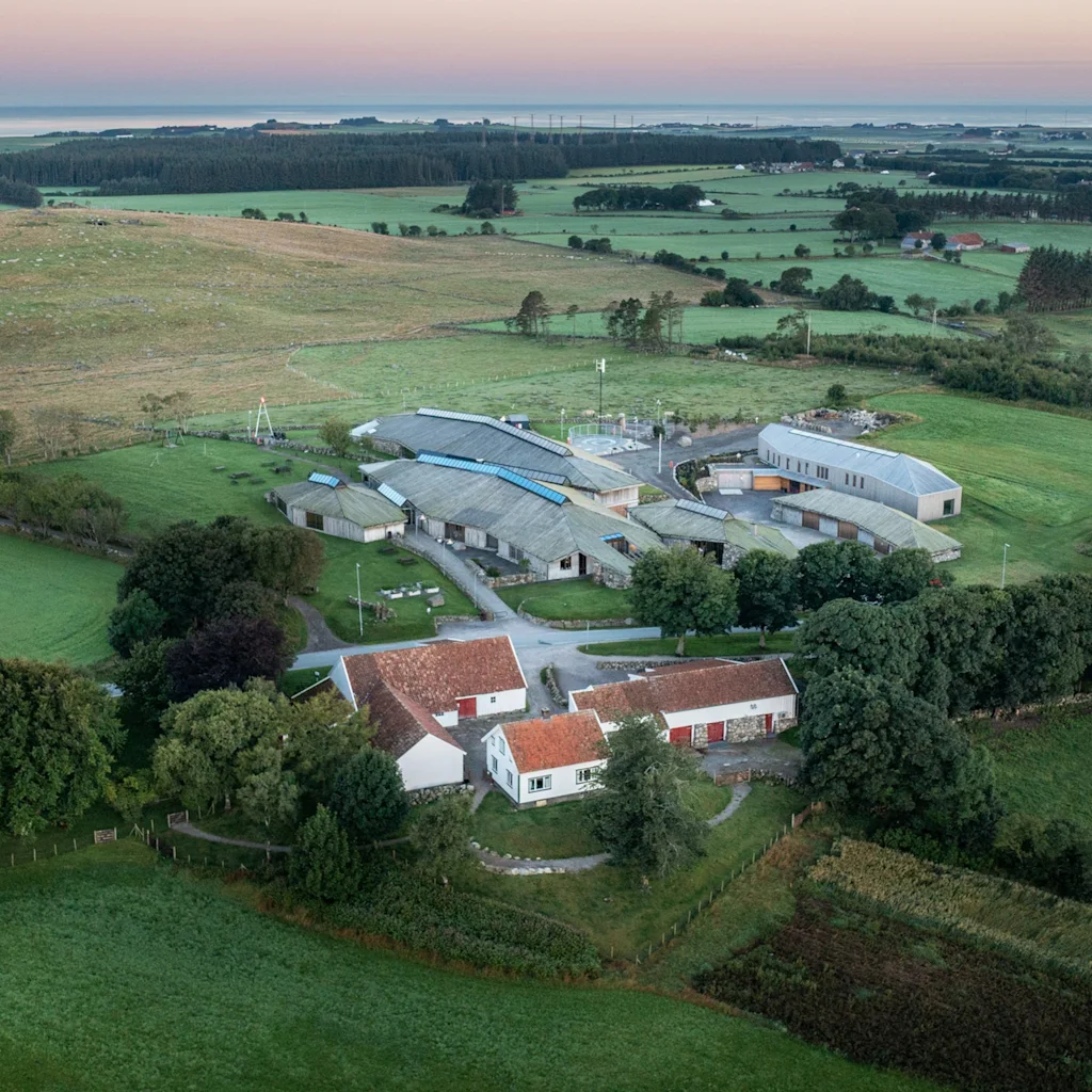 Ilmakuva maatilasta, jossa on perinteisiä punakattoisia taloja ja moderneja rakennuksia rehevien vihreiden peltojen ja puiden ympäröimänä.