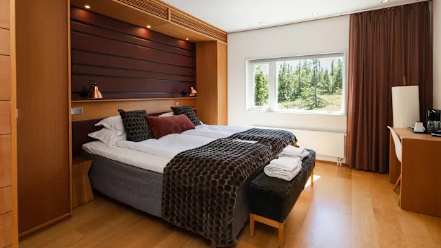 A neatly made bed in a bright, wood-paneled bedroom with a window showing a forest view.