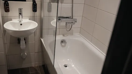 Puhdas, moderni kylpyhuone, jossa on valkoinen pesuallas ja kylpyamme lasisella suihkuseinällä. Annostelijat on asennettu seinälle.