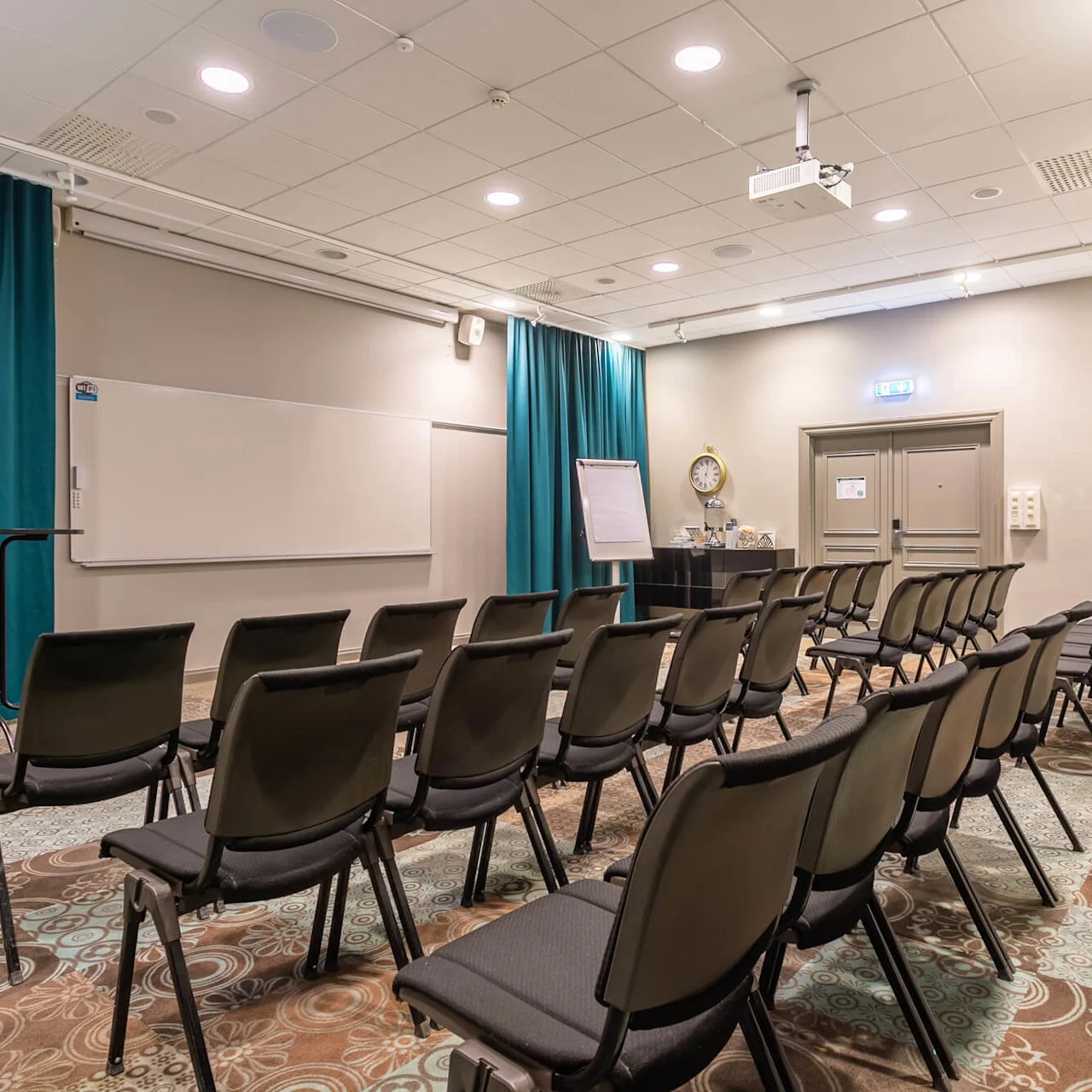 Rækker af tomme stole vender mod et whiteboard og en flipover i et tæppebelagt konferencerum med turkise gardiner og loftmonteret projektor, hvilket skaber en formel mødeindstilling.