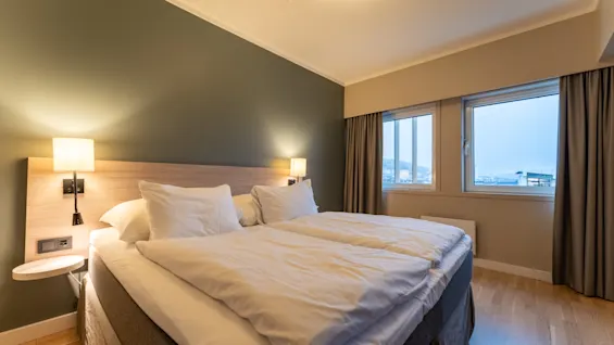 Et koselig hotellsoverom med en stor seng, nattbordslamper og vinduer som tilbyr utsikt over utsiden.