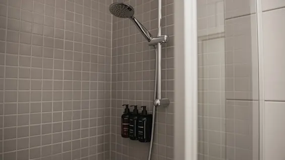 Showerhead affixed to a tiled bathroom wall with three bottles of toiletries on a shelf. The tiles are light gray, creating a simple and clean environment.