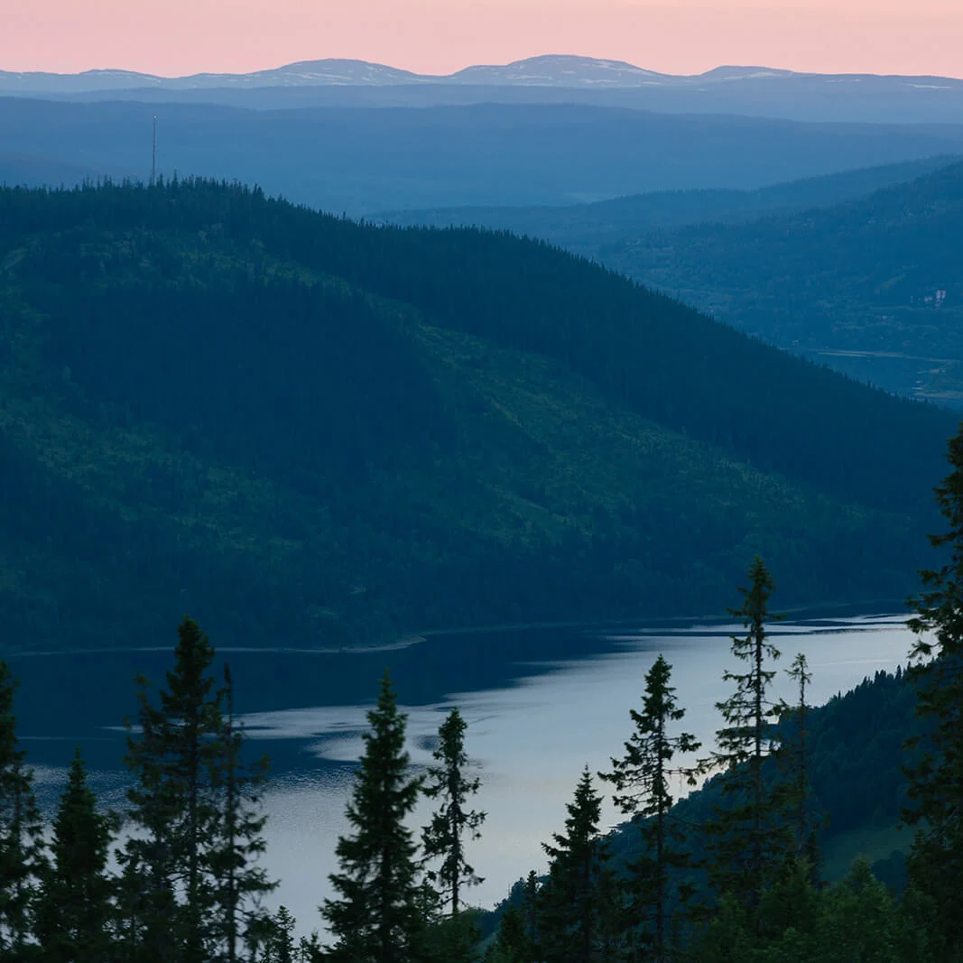 Layered mountains at sunset with a lake, silhouette of conifer trees in the foreground.
