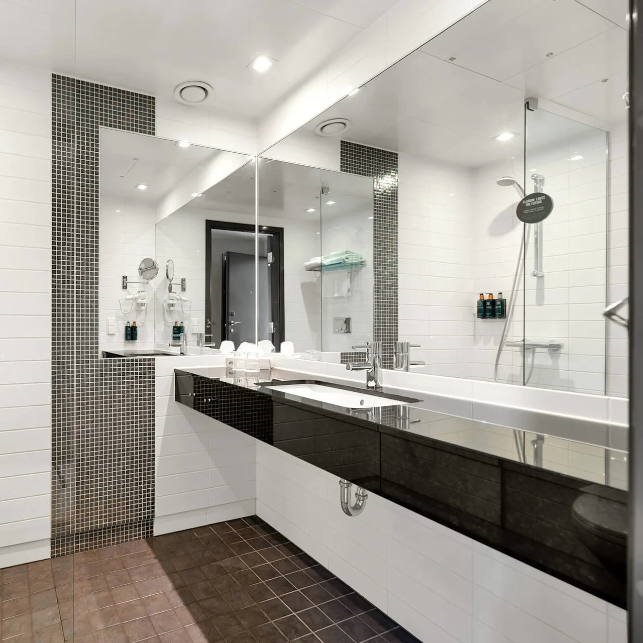 Shower with glass enclosure and black tiles, beside a large mirror and wide sink on a dark countertop. Bright lighting and white tiled walls create a modern bathroom atmosphere.