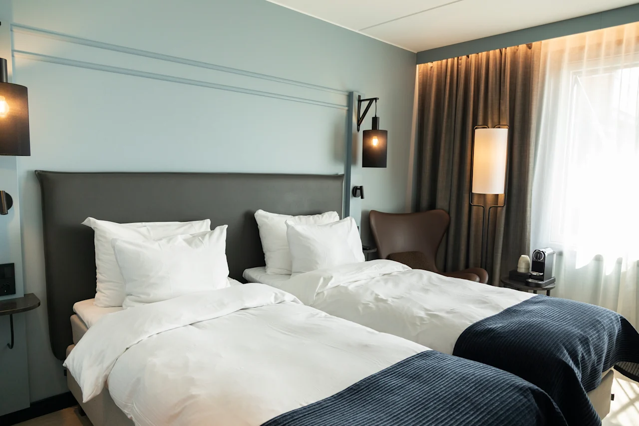 Et stilfullt hotellrom med to senger, skarpe hvite sengetøy, blå pledd og en lyseblå vegg. Myk belysning og en kaffemaskin.