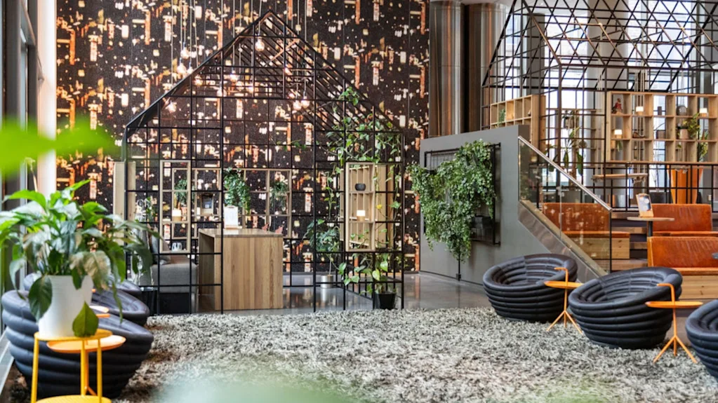 Et moderne kontorlokale med geometriske metalstrukturer, der ligner huse, omgivet af grønne planter og moderne møbler, mod en væg med bymønster.