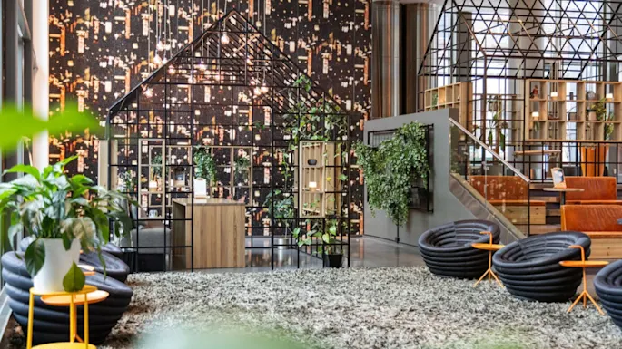 A modern office space features geometric metal structures resembling houses, with surrounding green plants and contemporary furniture, set against a wall with a cityscape pattern.