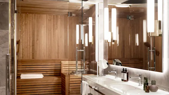 En badstue i tre med glassdører ligger ved siden av et moderne bad med dobbel servant, stort speil og stilig belysning. Håndklær og toalettartikler er pent ordnet.