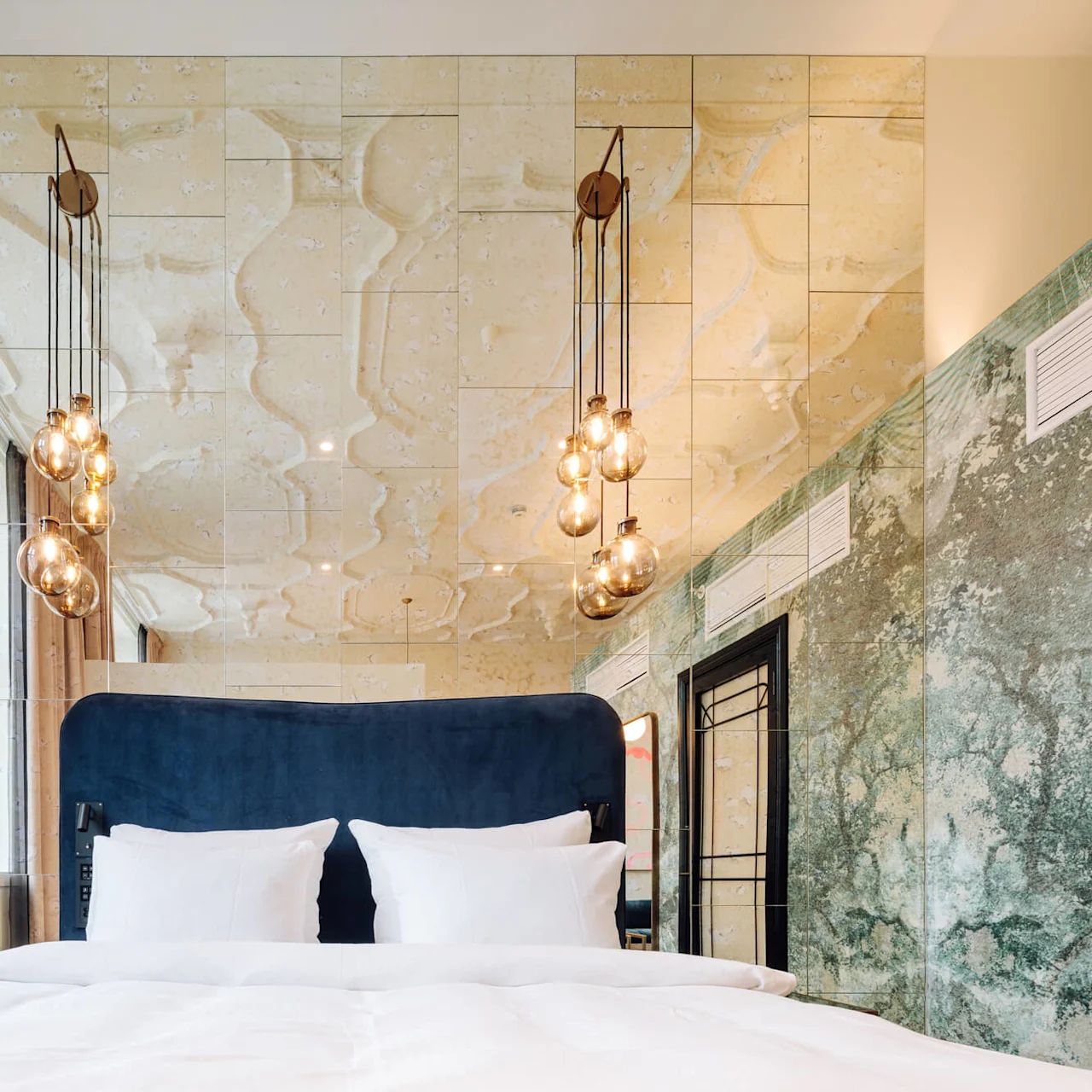 Bed with white linens, standing beneath hanging glass light fixtures. Mirrors and marbled walls surround the cozy modern room, with large curtains framing a window.