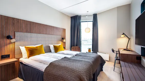Et pent arrangert hotellrom med dobbeltseng med gule puter, et trepanel, sengebordlamper, et skrivebord med lampe og et stort vindu med gardiner.
