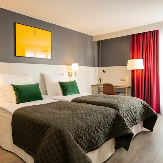 Två prydligt bäddade enkelsängar med gröna kuddar och grå täcken står i ett modernt hotellrum. Gul konst med texten "BANANA" hänger ovanför; ett skrivbord och stol syns nära röda gardiner.