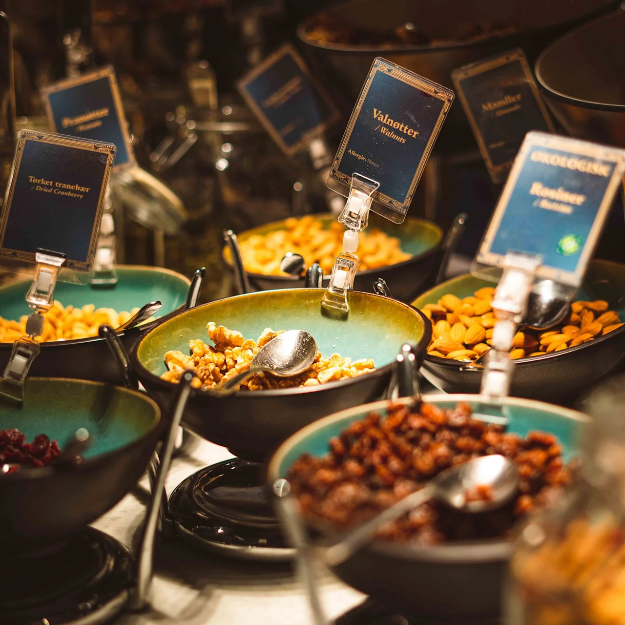 Bowls of assorted nuts and dried fruits are arranged with spoons. Labels read: “Valnötter, Mandlar, Torkat tranbär, Ekologisk russin.” They are on a buffet-style setup.