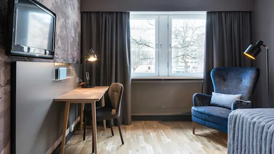 Ett rum har en väggmonterad TV, ett skrivbord i trä med en tänd lampa och en mörkblå fåtölj bredvid ett stort fönster med stängda gardiner.