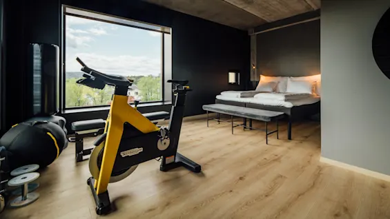 En gul motionscykel står stille i et moderne soveværelse med dobbeltseng og stort vindue. Værelset har minimalistiske møbler og trægulv. Tekst: "TECHNOGYM".
