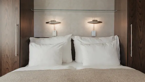 En prydligt bäddad hotellsäng med vita kuddar och en mysig filt, flankerad av träpaneler och upplyst av varma vägglampor.