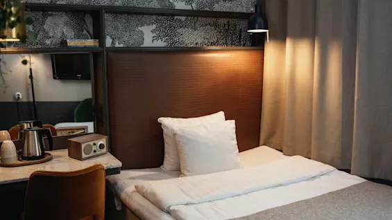 Et hyggeligt hotelværelse med en seng, varm belysning og et lille skrivebordsområde med en elkedel og radio, der indbyder til afslapning.
