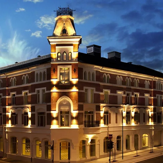 A multi-story building with arched windows is illuminated at night. It stands on a street corner, surrounded by similar architecture, under a twilight blue sky.