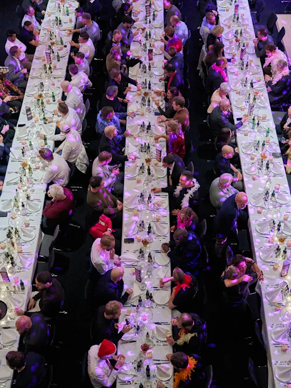 En stor grupp människor samlade runt långa bord, som njuter av ett socialt evenemang eller middag med festliga dekorationer.