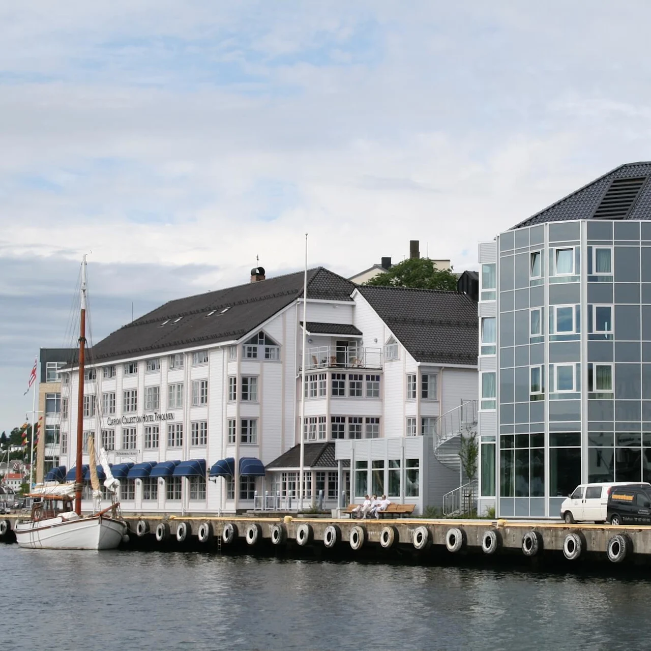 Et hotell ved vannet med en tradisjonell fasade og en moderne glassbygning står ved en brygge. Flere biler og en fortøyd seilbåt er til stede. Tekst: "Clarion Collection Hotel Tyholmen."