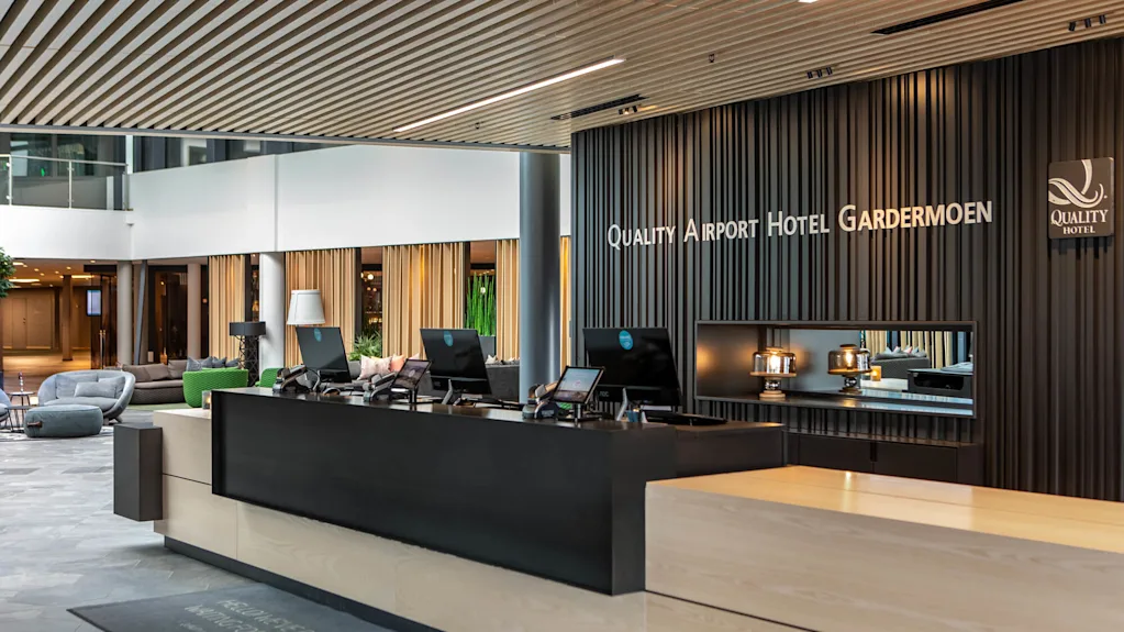 Resepsjonsdisk med datamaskiner i en moderne hotellobby, med elegant design og merkevarebygging. Tekst på veggen lyder "Quality Airport Hotel Gardermoen."