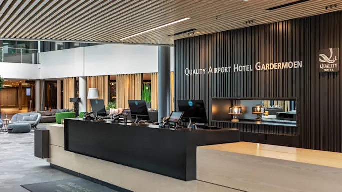 Reception desk with computers in a modern hotel lobby, featuring sleek design and branding. Text on the wall reads "Quality Airport Hotel Gardermoen."